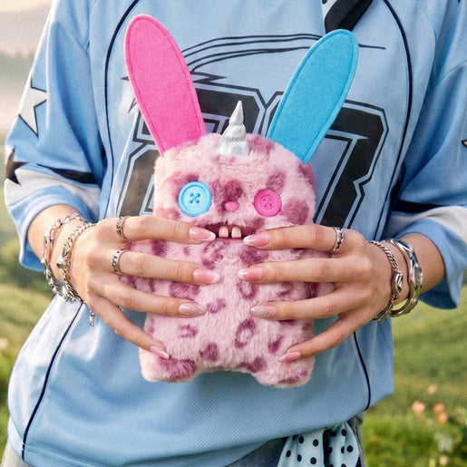 Person holding a Fuggler Fugglercorns Rabid Rabbit plush toy with pink and dark purple spotted fur, tall ears with blue and pink insides, a silver unicorn horn, mismatched button eyes, and two large buck teeth.