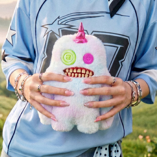 Person holding a Fuggler Fugglercorns Sasquoosh plush toy with pastel ombre fluff, a metallic bright pink unicorn horn, mismatched green and pink button eyes, and a wide mouth full of human-like teeth.