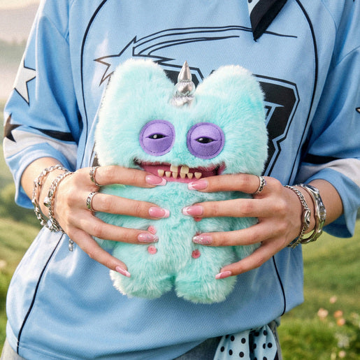 Person holding a Fuggler Fugglercorns Stinkface plush toy with fluffy pastel blue fur, a shiny silver unicorn horn, purple sleepy eyes, human-like teeth, and four pink buttons on its belly.