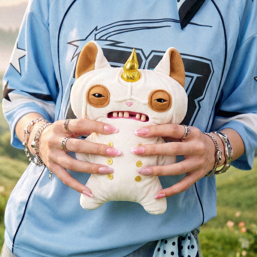 Person holding a Fuggler Fugglercorns Wrinkle McStinkles plush toy with wrinkled white fur, a metallic gold unicorn horn, sleepy brown eyes, human-like teeth, and six gold buttons on its belly.
