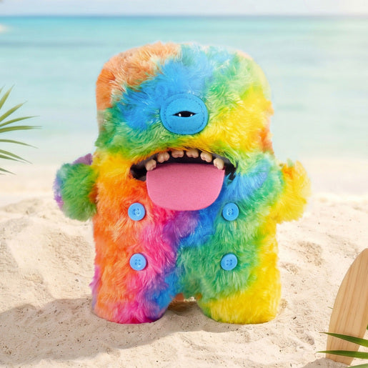 Fuggler Nipple Fuggs Oogah Boogah plush toy with fluffy rainbow tie-dye fur, a single blue eye, human-like teeth, a large pink tongue, and four blue buttons on its belly on a sandy beach