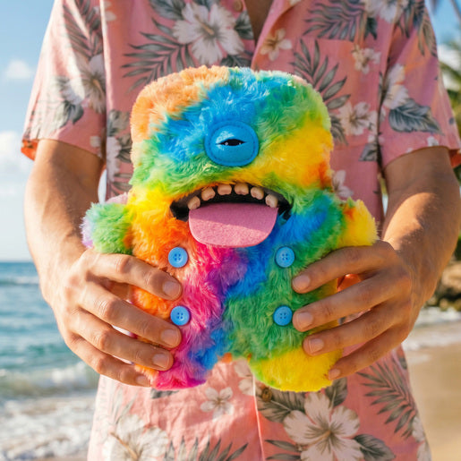 Person holding a Fuggler Nipple Fuggs Oogah Boogah plush toy with fluffy rainbow tie-dye fur, a single blue eye, human-like teeth, a large pink tongue, and four blue buttons on its belly.