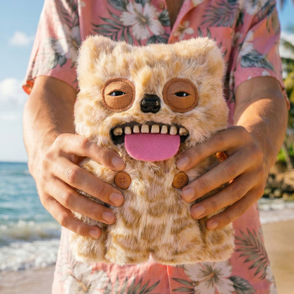 Person holding a Fuggler Nipple Fuggs Sir Belch plush toy in fluffy tan and brown spotted fur with human-like teeth, a pink tongue sticking out, a black nose, sleepy eyes, and two brown buttons on its belly.