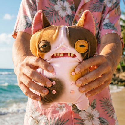Person holding a Fuggler Nipple Fuggs Wrinkle McStinkles plush toy with pink and brown patches, human-like teeth, sleepy eyes, and four pink buttons on its bellywith a beach in the background
