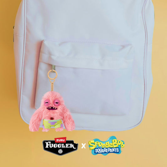 Pink plush keychain with a yellow tag attached to a white backpack on a yellow background, featuring Fuggler and SpongeBob SquarePants logos.