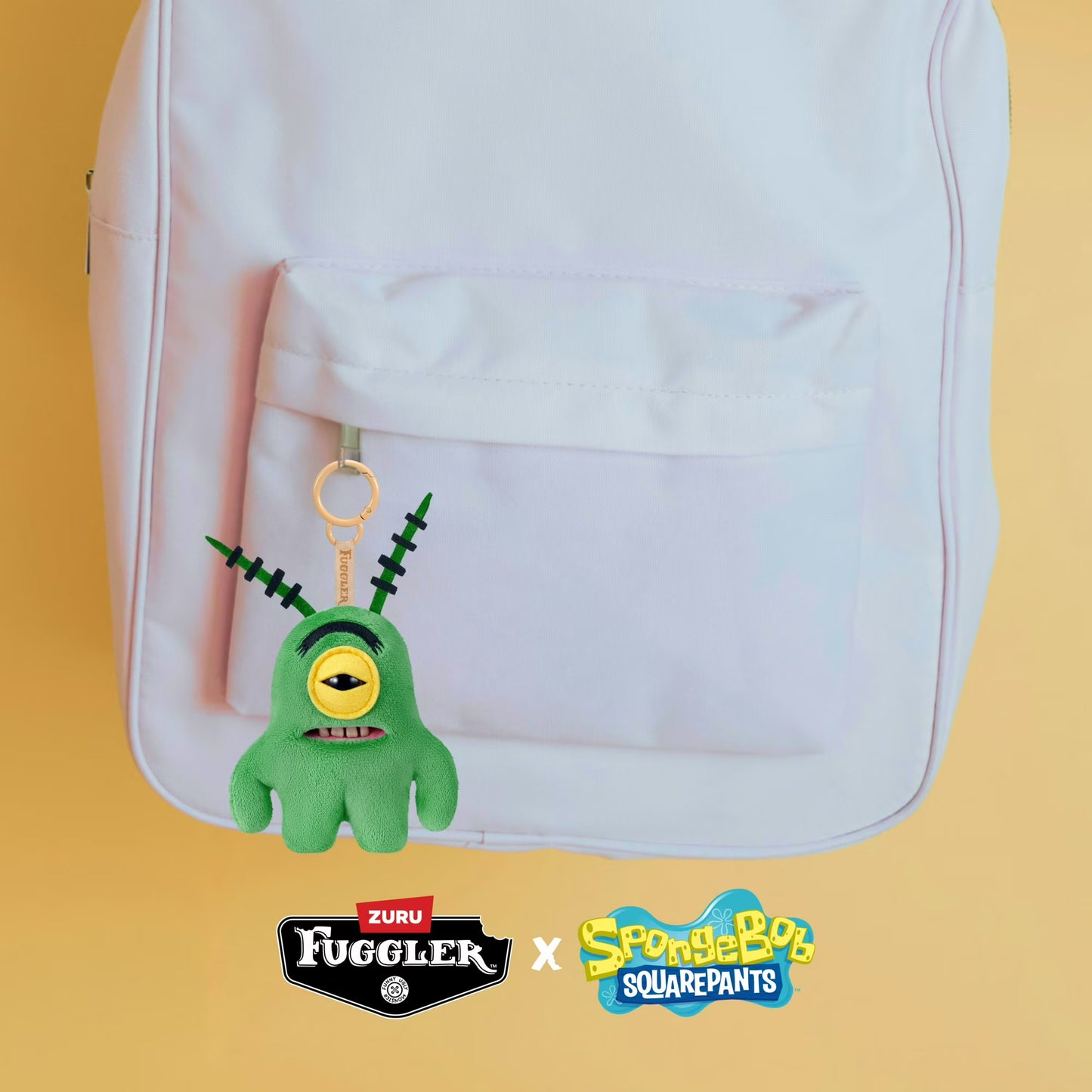 Green plush keychain with a yellow eye and black mouth attached to a white backpack on a yellow background, featuring Zuru Fuggler and SpongeBob SquarePants logos.