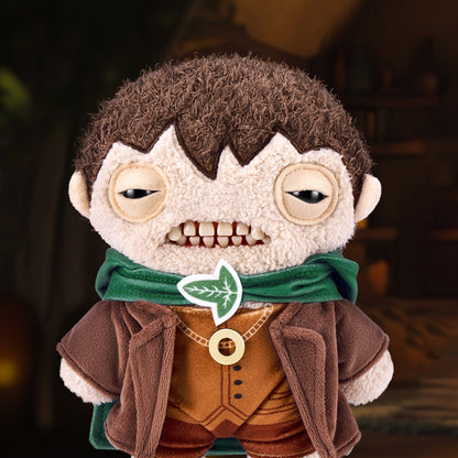 The Frodo Baggins Fuggler plush toy displayed against a warm Hobbit-hole background, emphasizing his tan fleece fur and mischievous expression.