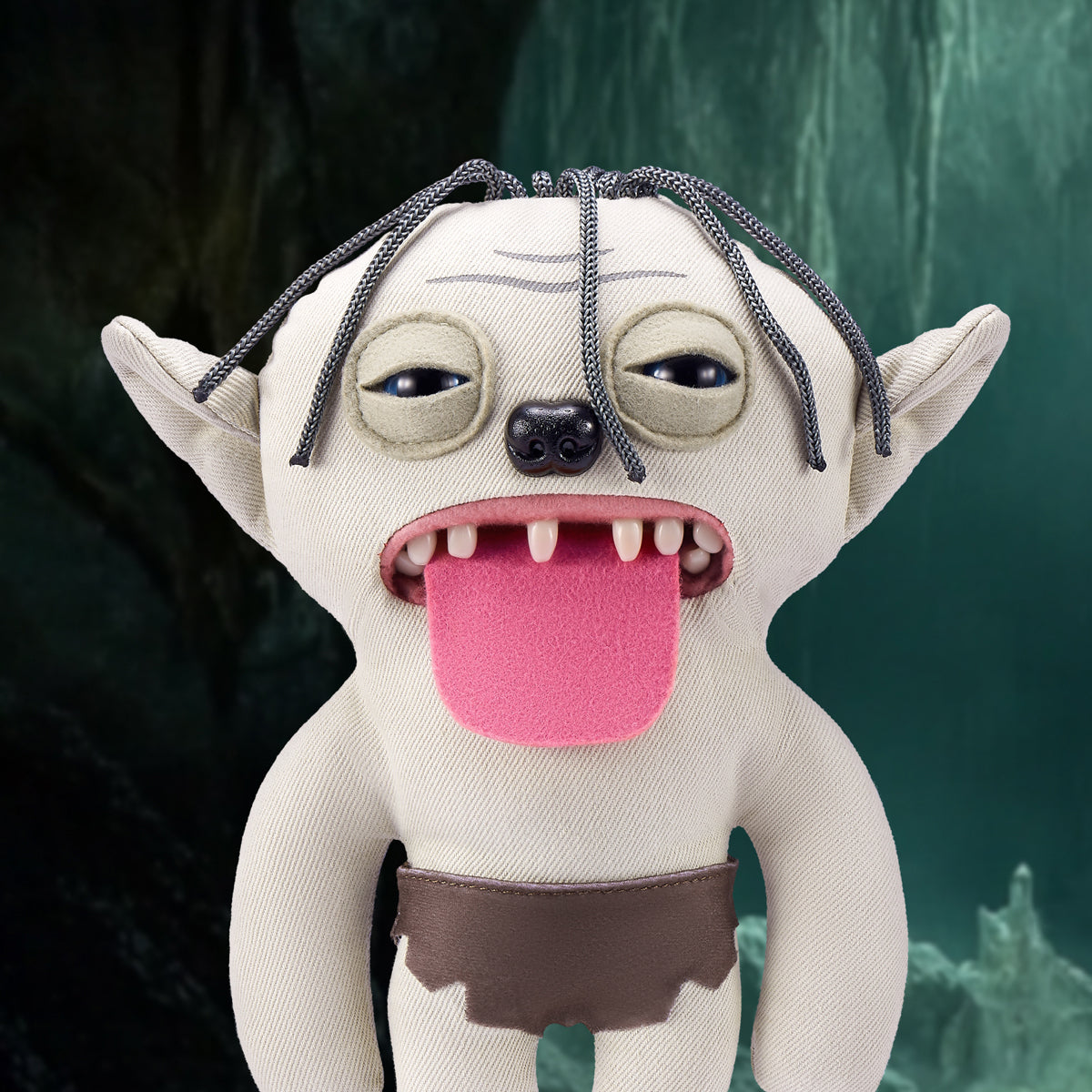 The Gollum Fuggler plush toy displayed against a dark, cave-like background, emphasizing his beige fur, large ears, and creepy expression.