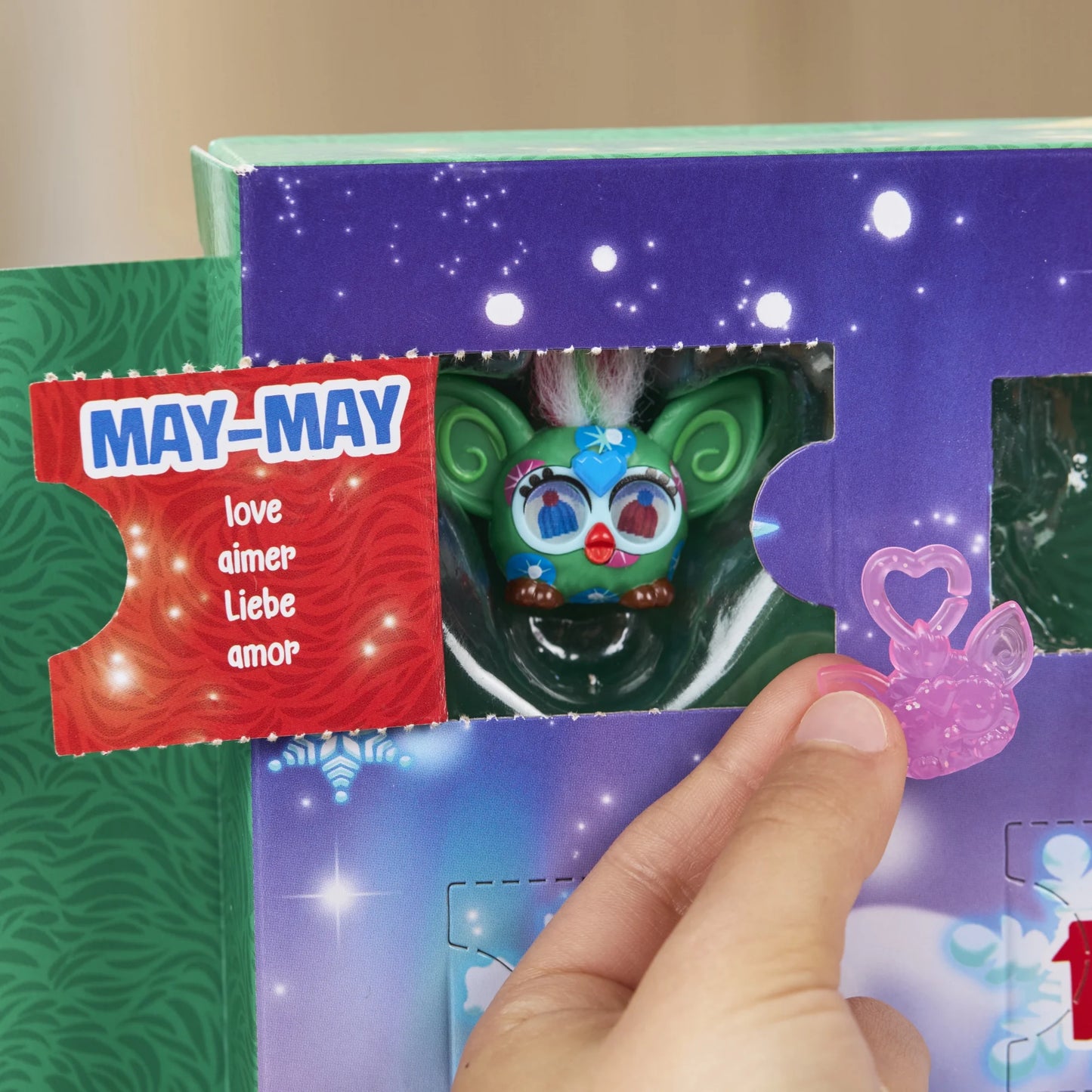A hand holding a furby toy from the  Furby Minis Advent Calendar box with an open tab containing a green holiday furby and the words "May - love" on the inside of the tab.