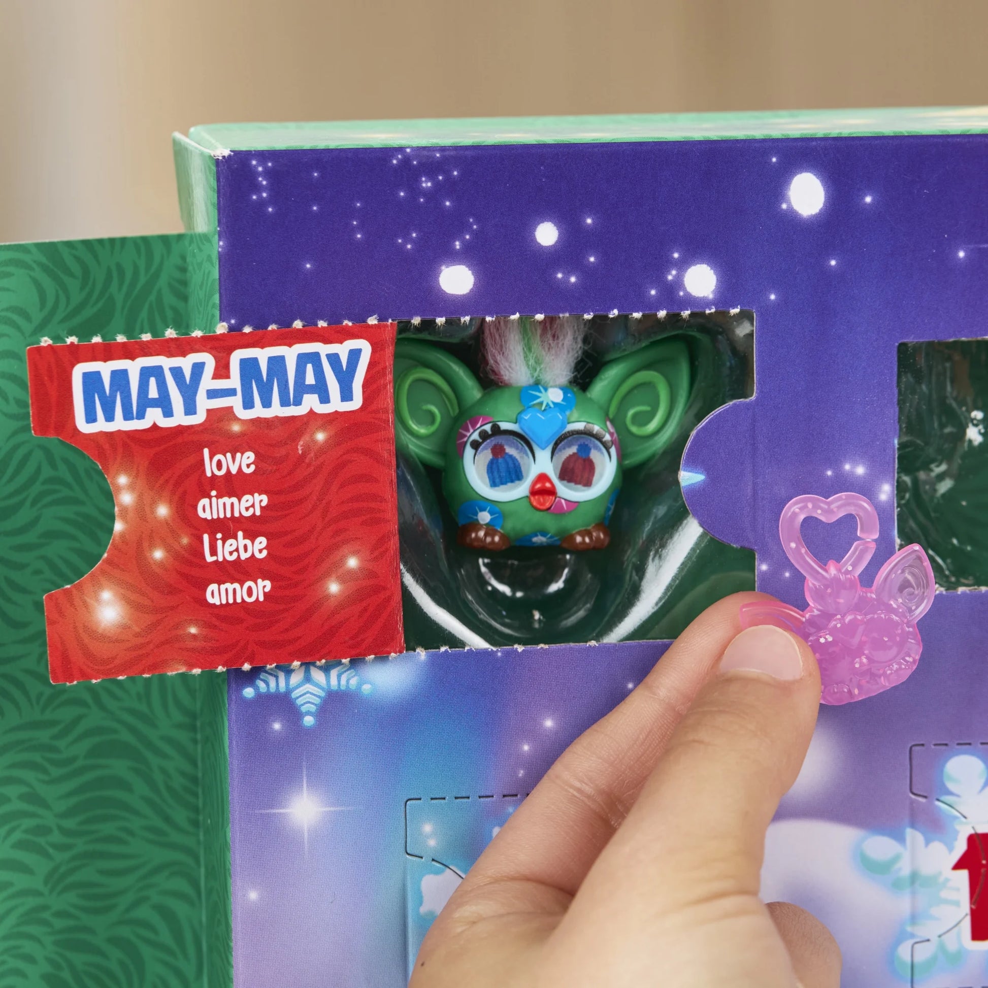 A hand holding a furby toy from the  Furby Minis Advent Calendar box with an open tab containing a green holiday furby and the words "May - love" on the inside of the tab.