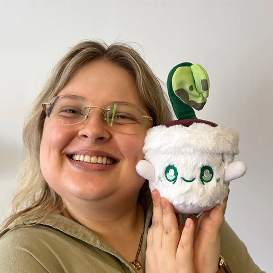 Person holding the Squishable Alter Egos Series 16 Plant Ghost 7-Inch Plush, featuring a round white ghost-like body sitting in a fuzzy white plush pot with a smiling face, green eyes, and a green plant sprout with leaves on its head.