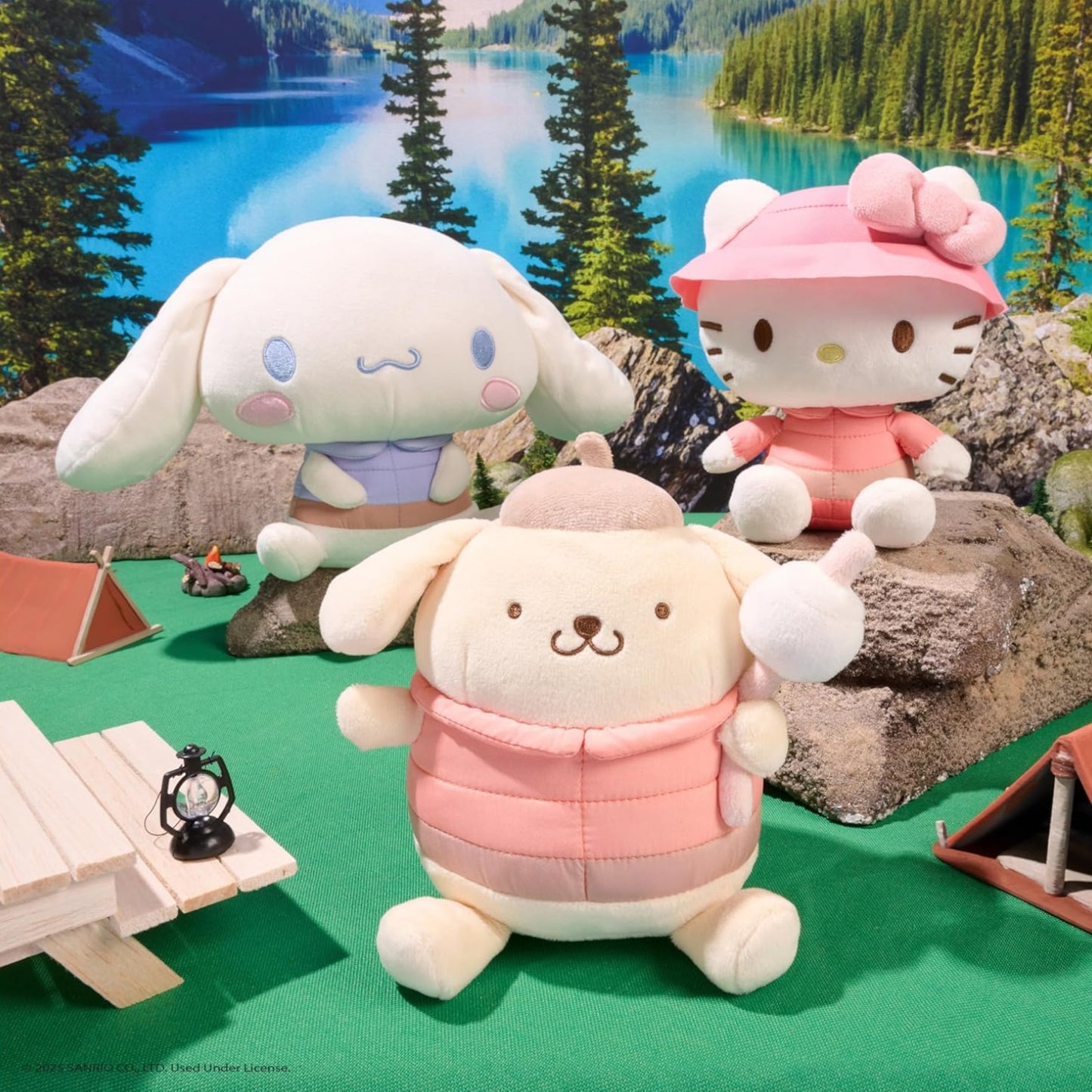 Three plush toys in a camping scene with a lake and trees in the background