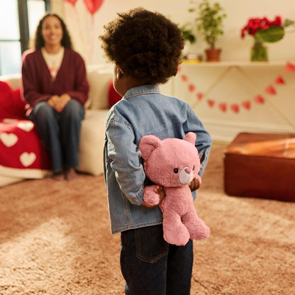 Child holding a pink teddy bear in a living room with a woman sitting on a couch.