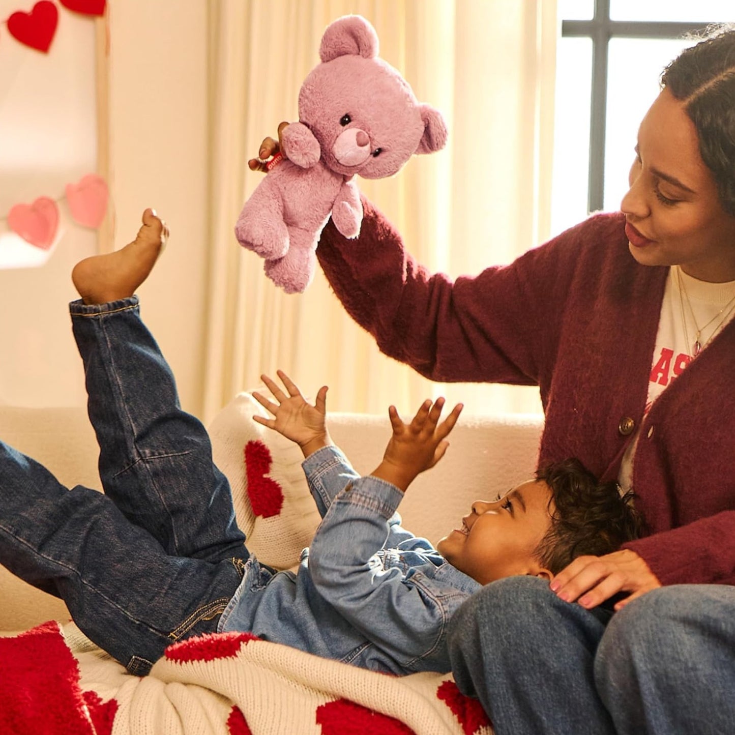 Woman playing with a child on a couch, holding a pink teddy bear with hearts in the background.