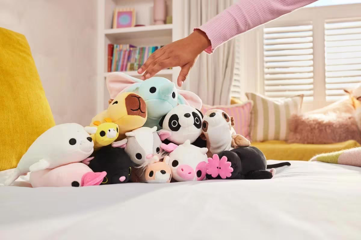 Hand reaching towards a collection of colorful plush toys on a bed.