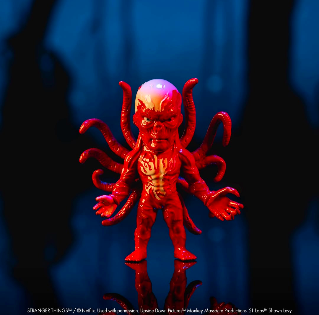 A red and orange horror character figurine with multiple tentacle-like arms and a glowing head, standing on a reflective surface against a dark blue-black background.