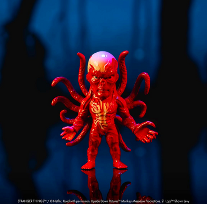 A red and orange horror character figurine with multiple tentacle-like arms and a glowing head, standing on a reflective surface against a dark blue-black background.