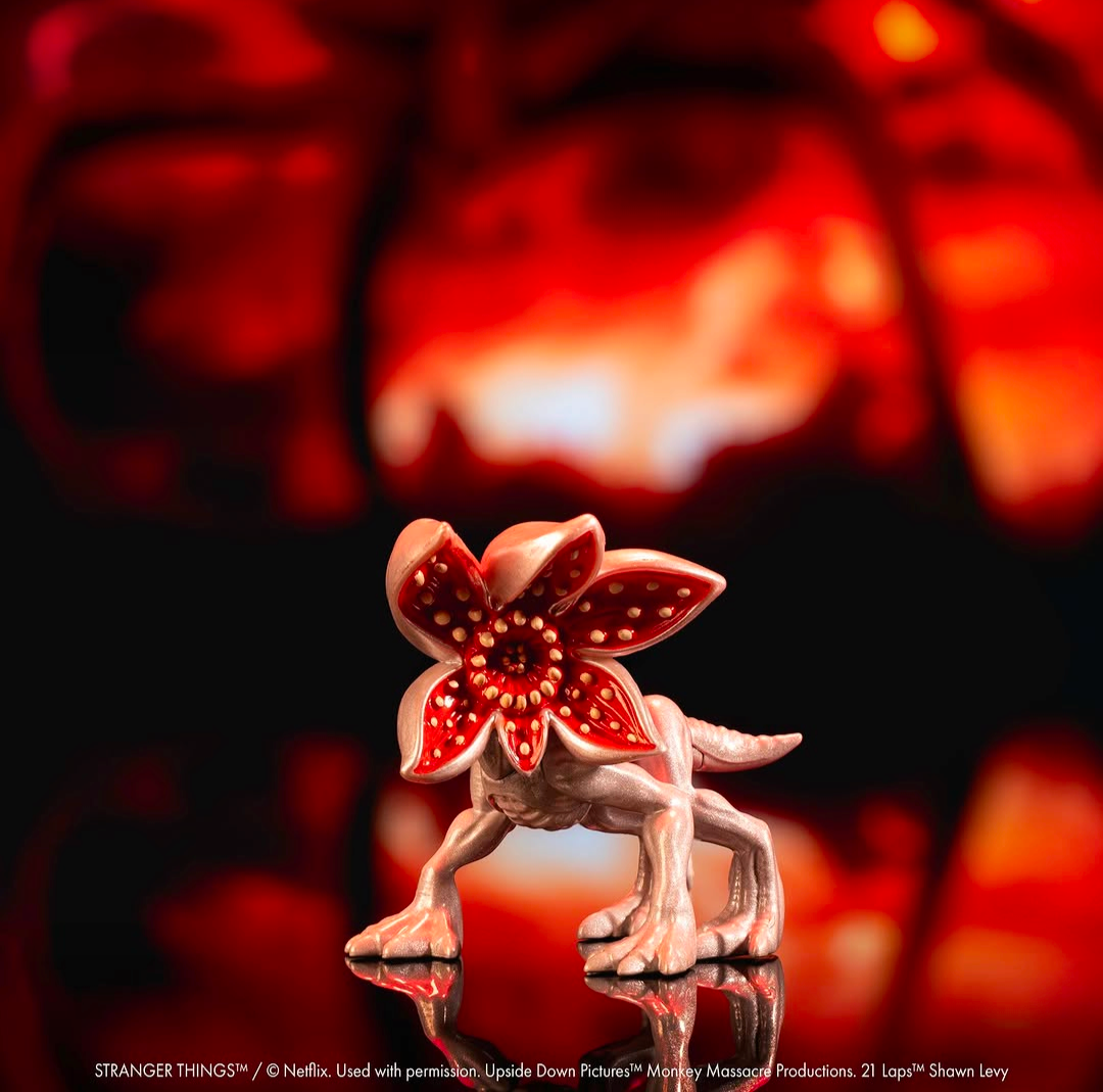 A small red and beige flower-like creature figurine stands on a reflective surface against a dark background with a blurred red abstract shape behind it.