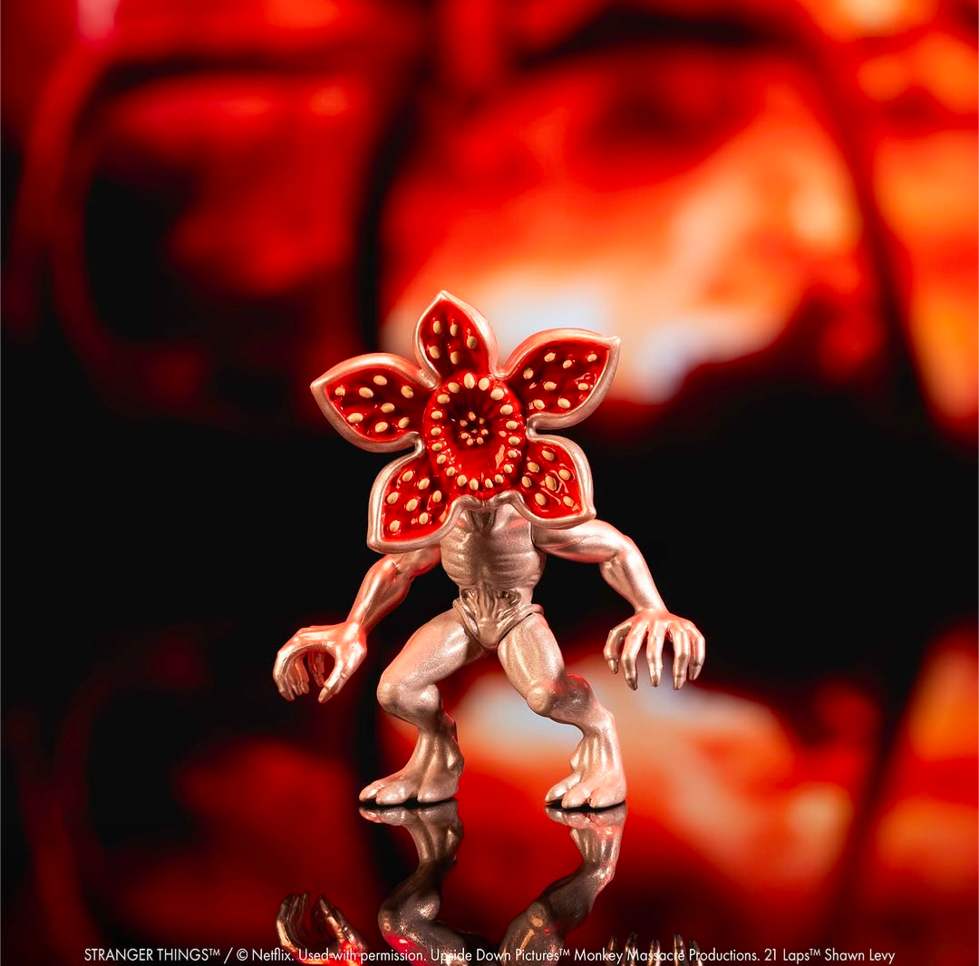 A small Stranger Things figurine of the Siren character, featuring a metallic silver body and a red flower-shaped head with yellow details, set against a dark background with a blurred red and black version of the character.