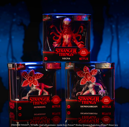 Three Stranger Things die-cast metal figures in retail packaging, arranged against a dark blue forest background. The top figure is Vecna, red, and the bottom two are Demogorgon and Demodog, both with glowing red flower-like heads and pale bodies.