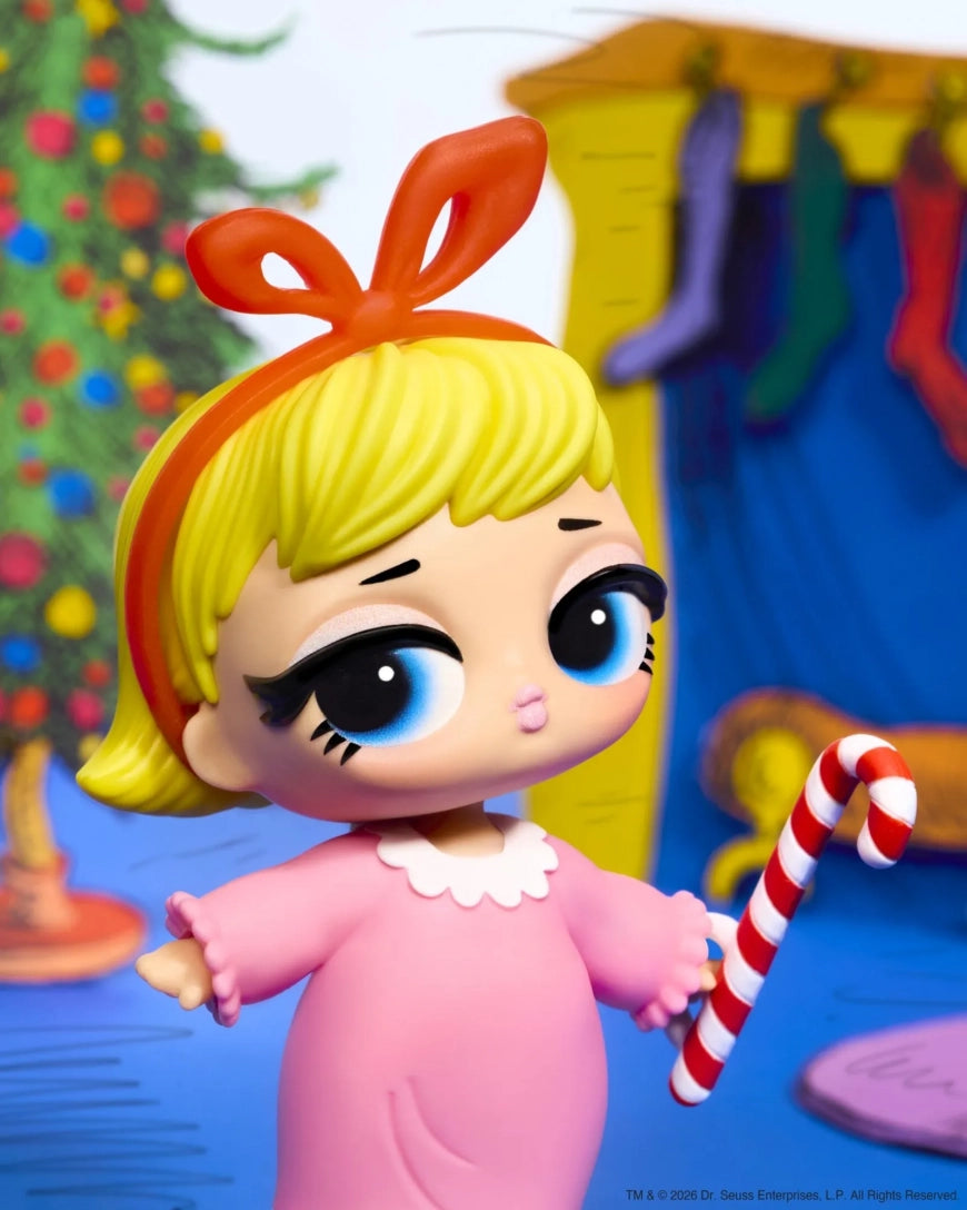 A stylized collectible doll with large blue eyes, yellow hair, and an orange bow, wearing a pink outfit and holding a red-and-white striped candy cane. The background is a colorful illustrated holiday scene with stockings and a tree.