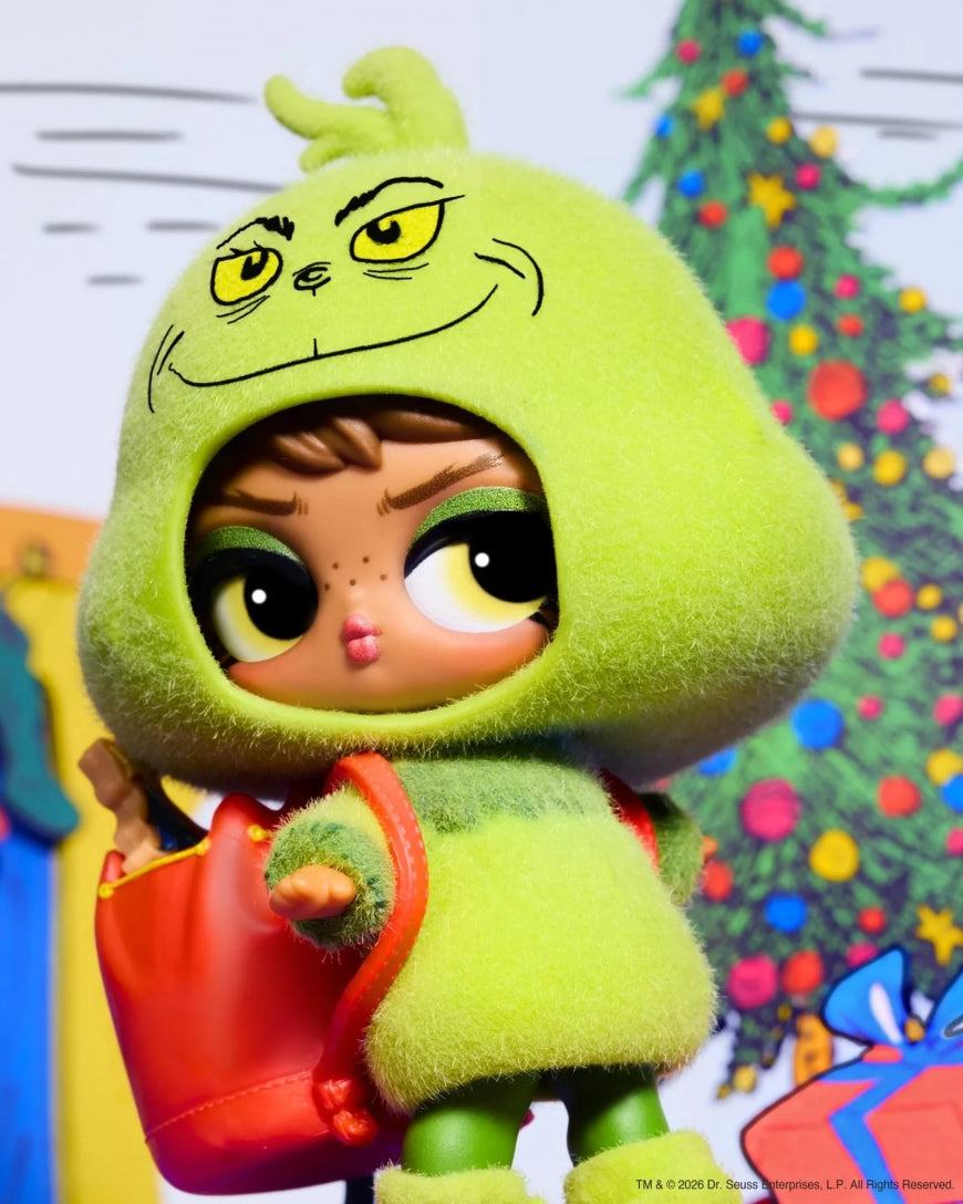 A collectible doll wearing a fuzzy lime-green Grinch-style hooded outfit with a red bag, posed in front of a decorated Christmas tree background.