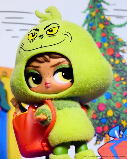 A collectible doll wearing a fuzzy lime-green Grinch-style hooded outfit with a red bag, posed in front of a decorated Christmas tree background.