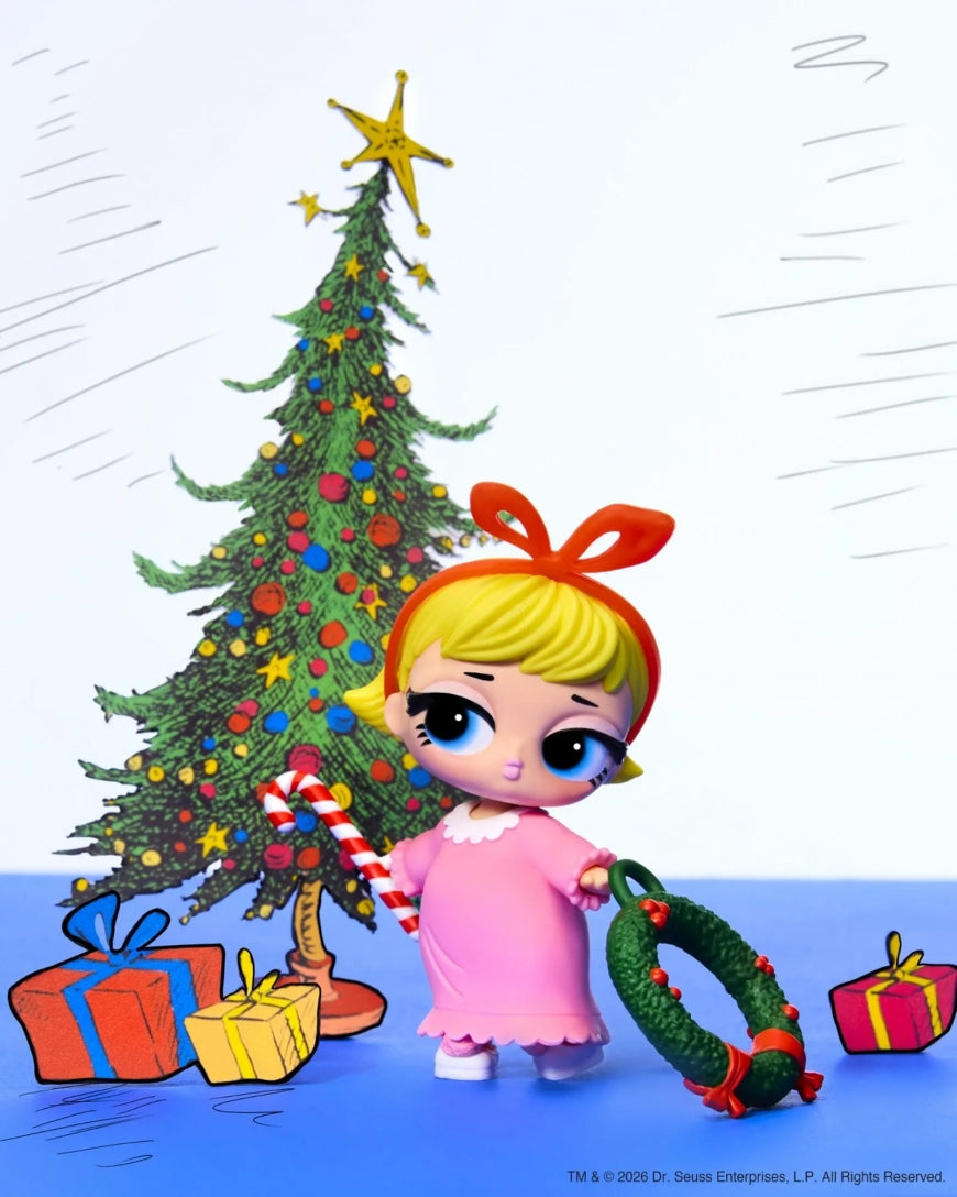 A stylized illustration of a blonde doll with a red bow headband, wearing a pink dress, standing in front of a decorated Christmas tree and holding a candy cane and a green wreath, with wrapped gifts on the floor.