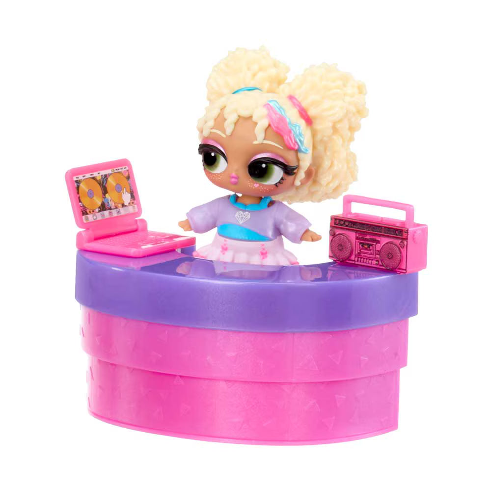 A LOL Surprise! doll with blonde pigtails sits behind a pink and purple counter, with a pink laptop and boombox accessories on either side, on a white background.