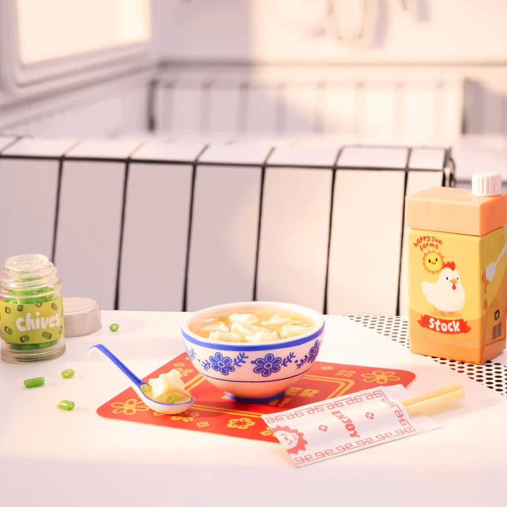 Bowl of soup with a spoon on a table next to a container labeled 'Chives' and a box labeled 'Stock'.