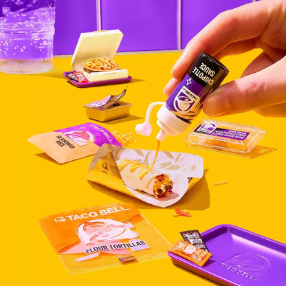 Person pouring Taco Bell sauce onto a tortilla with various Taco Bell products on a yellow table.