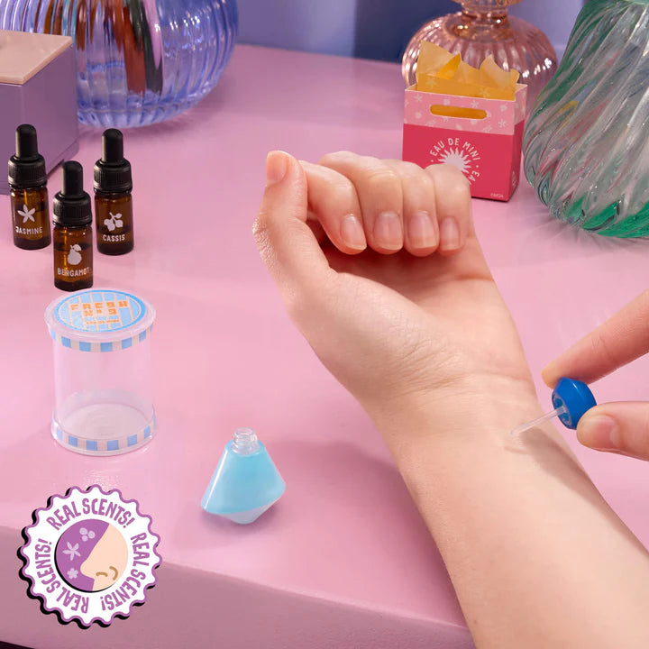 A hand applying a drop of liquid from a blue miniature perfume bottle onto a wrist, next to a "Real Scents!" badge indicating the resin is actually scented.
