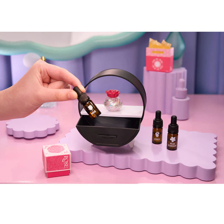 A hand placing a small brown oil bottle into the drawer of a black circular display stand, which features a finished pink rose-shaped perfume bottle sitting on the top shelf.