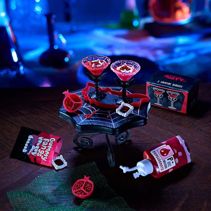 Gory Halloween cocktail set with two red blood-stained martini glasses on a spiderweb tray, plus fake blood tubes, a pomegranate prop, and a branded product box on a dark tabletop with a blurred blue bar background.