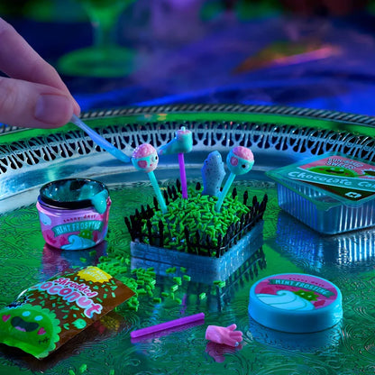 A hand is placing a blue spatula into a miniature slime garden kit on a reflective green tray under blacklight. The kit contains various colorful slime ingredients and finished miniatures, including a pink and green slime lollipop, a blue slime egg, and a small pink slime hand.