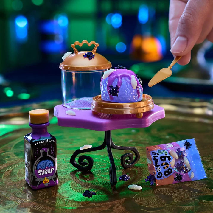 A Halloween-themed miniature toy setup on a reflective green surface, featuring a small purple slime-like dessert on a gold stand, a purple bottle labeled 'Lemon Drank Syrup,' a purple bag labeled 'Spooky Bag,' and a hand holding a small spatula.
