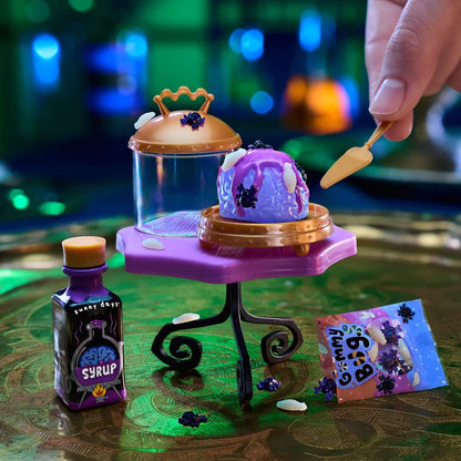 A Halloween-themed miniature toy setup on a reflective green surface, featuring a small purple slime-like dessert on a gold stand, a purple bottle labeled 'Lemon Drank Syrup,' a purple bag labeled 'Spooky Bag,' and a hand holding a small spatula.