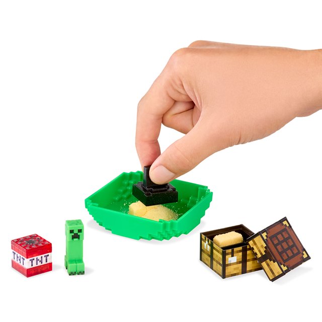 A hand using a stamp tool to pack yellow kinetic sand "gunpowder" into a miniature TNT block, displayed next to a green Creeper figure.