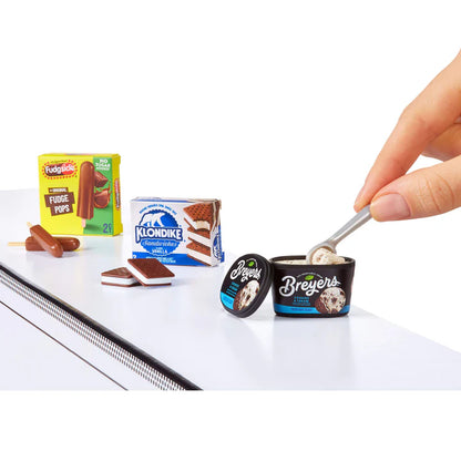 A hand using a silver spoon to scoop miniature Breyers Cookies & Cream ice cream, displayed next to boxes of Fudgsicle and Klondike Bar replicas.