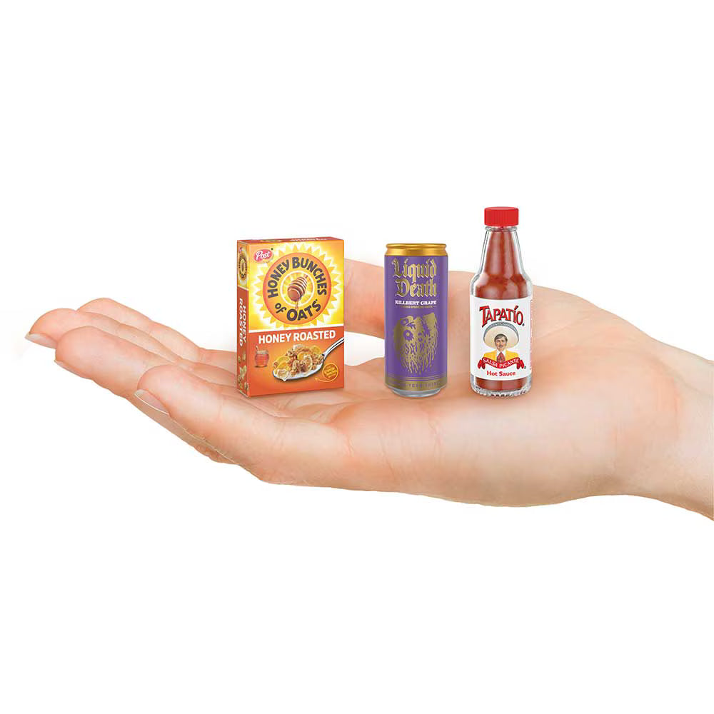 Three miniature toy food and drink containers displayed on an open hand against a plain white background: a Honey Burger Oats box, a Purple Drink can, and a Tapatio hot sauce bottle.