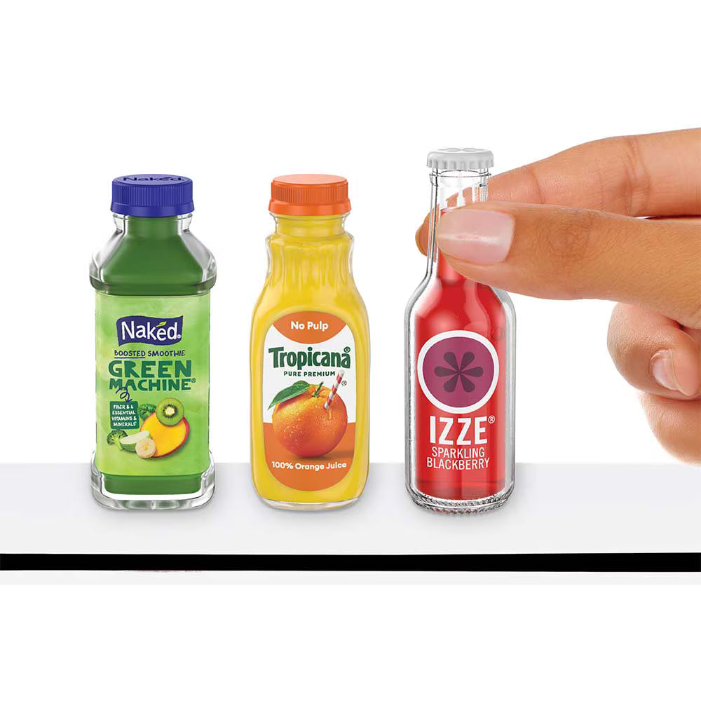 Three miniature juice bottles are lined up on a white surface: a green Naked Green Machine bottle, a Tropicana orange juice bottle, and a red IZZE sparkling blackberry bottle. A human hand is reaching in from the right to touch the IZZE bottle.