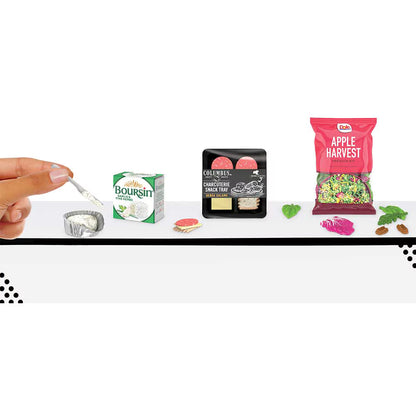 A miniature toy food playset on a white surface, featuring a hand holding a tiny spoon with white frosting, a small Boursin box, a chocolate chip cookie, a black bag of Oreo's, and a red Apple Harvest bag, all scaled down.