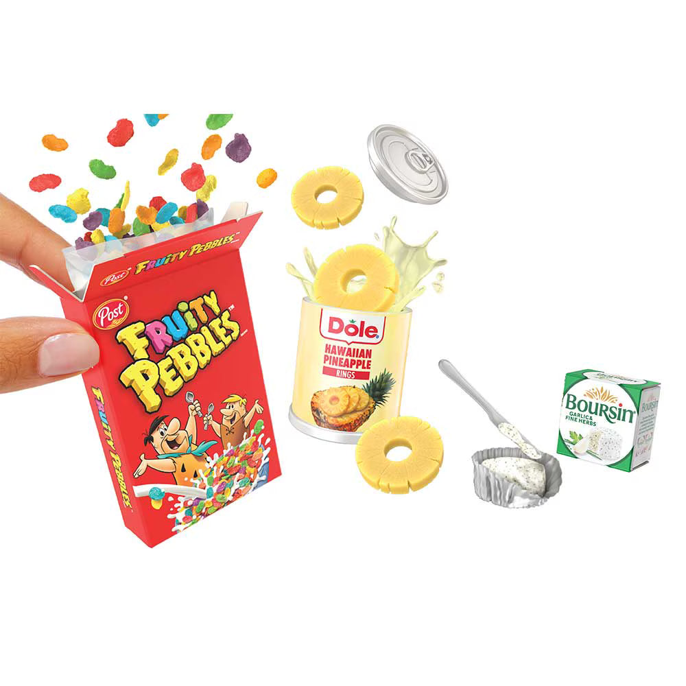 A miniature toy set depicting a small red box of Pez Fruity Pebbles being held by a hand, alongside a can of Dole Hawaiian Pineapple, loose yellow pineapple rings, colorful candy pieces, a silver foil cup, and a small Boursin cheese box, all on a white background.
