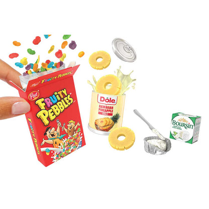 A miniature toy set depicting a small red box of Pez Fruity Pebbles being held by a hand, alongside a can of Dole Hawaiian Pineapple, loose yellow pineapple rings, colorful candy pieces, a silver foil cup, and a small Boursin cheese box, all on a white background.
