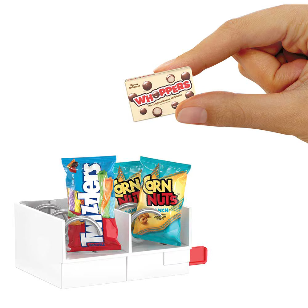 A hand holding a miniature Whoopsers candy box above a small white organizer filled with oversized candy bags, including Twix and Corn Nuts, on a white background.