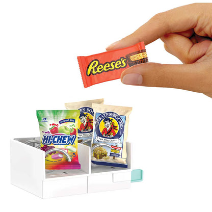 A hand holds a miniature orange Reese's candy above a small white organizer filled with tiny snack packages, including Hi-Chew and Warheads.