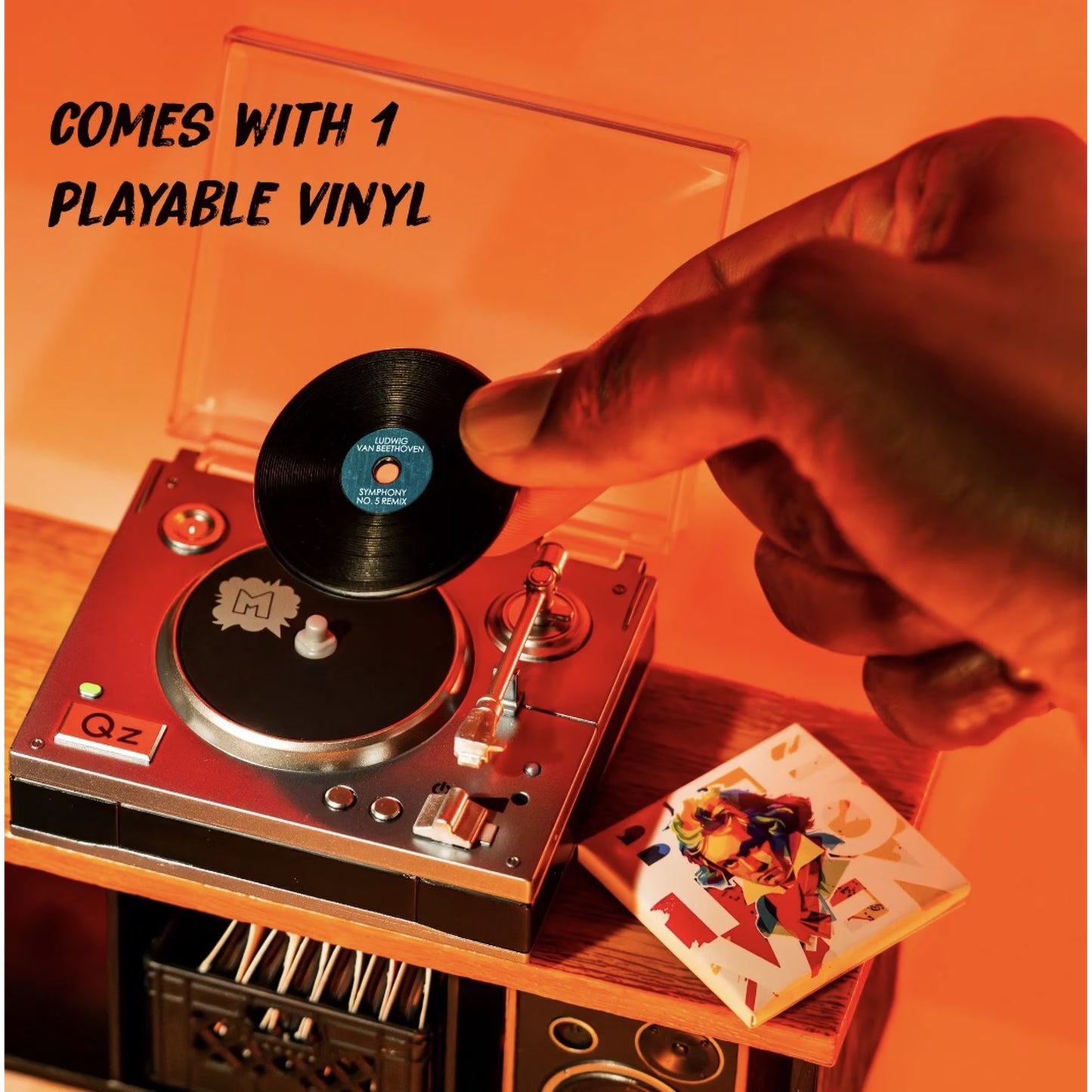 A hand placing a black vinyl record onto a turntable in a warm orange-lit setting, with text reading 'COMES WITH 1 PLAYABLE VINYL' and a small illustrated card beside the turntable.