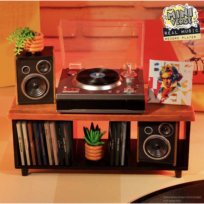 A miniature record player set displayed on a small wooden stand, featuring a turntable with a clear dust cover, two matching speakers, a vinyl record, a colorful album cover, and small potted plants. The scene is lit with warm orange lighting against a matching warm-toned background.