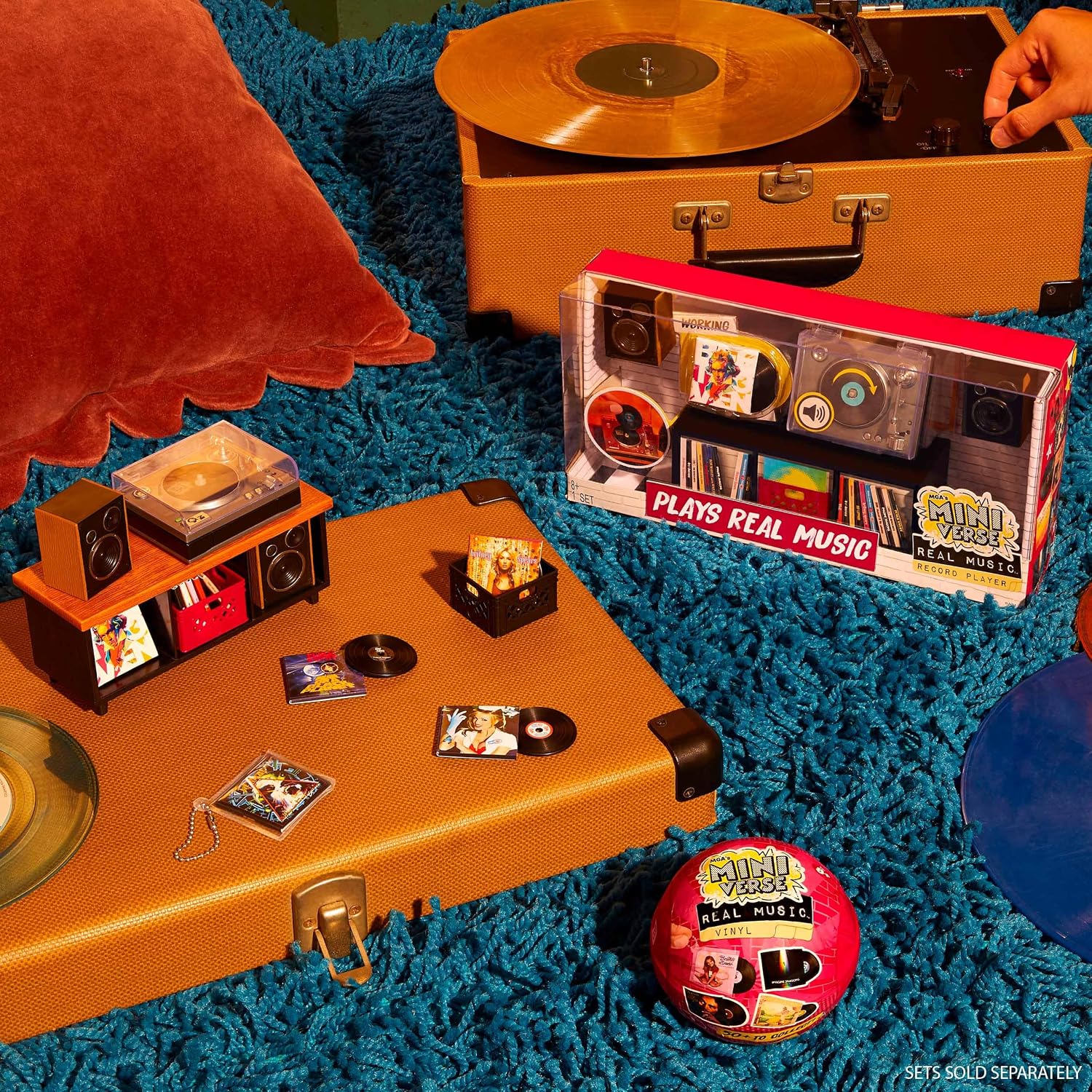 A styled product arrangement on a bright blue shag rug featuring a toy record player set in a clear suitcase, a smaller orange record player with accessories, a vinyl record, and a pink ball-shaped item labeled 'Play with Me' and 'Real Music.' A hand is visible at the top right near the larger record player.