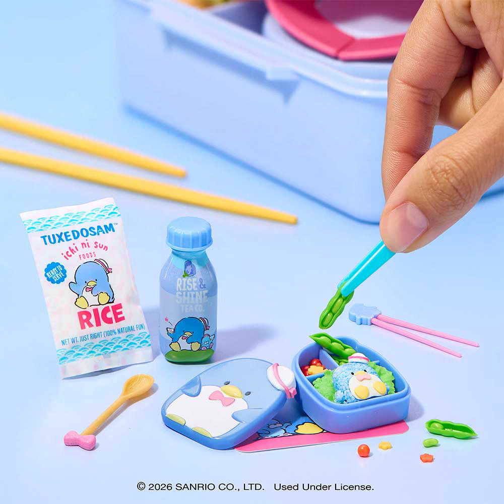 A miniature cartoon-themed lunch set with a blue bento box, small bottle, rice packet, chopsticks, spoon, and accessories, shown on a light blue surface with a hand placing a small green accessory into the box.